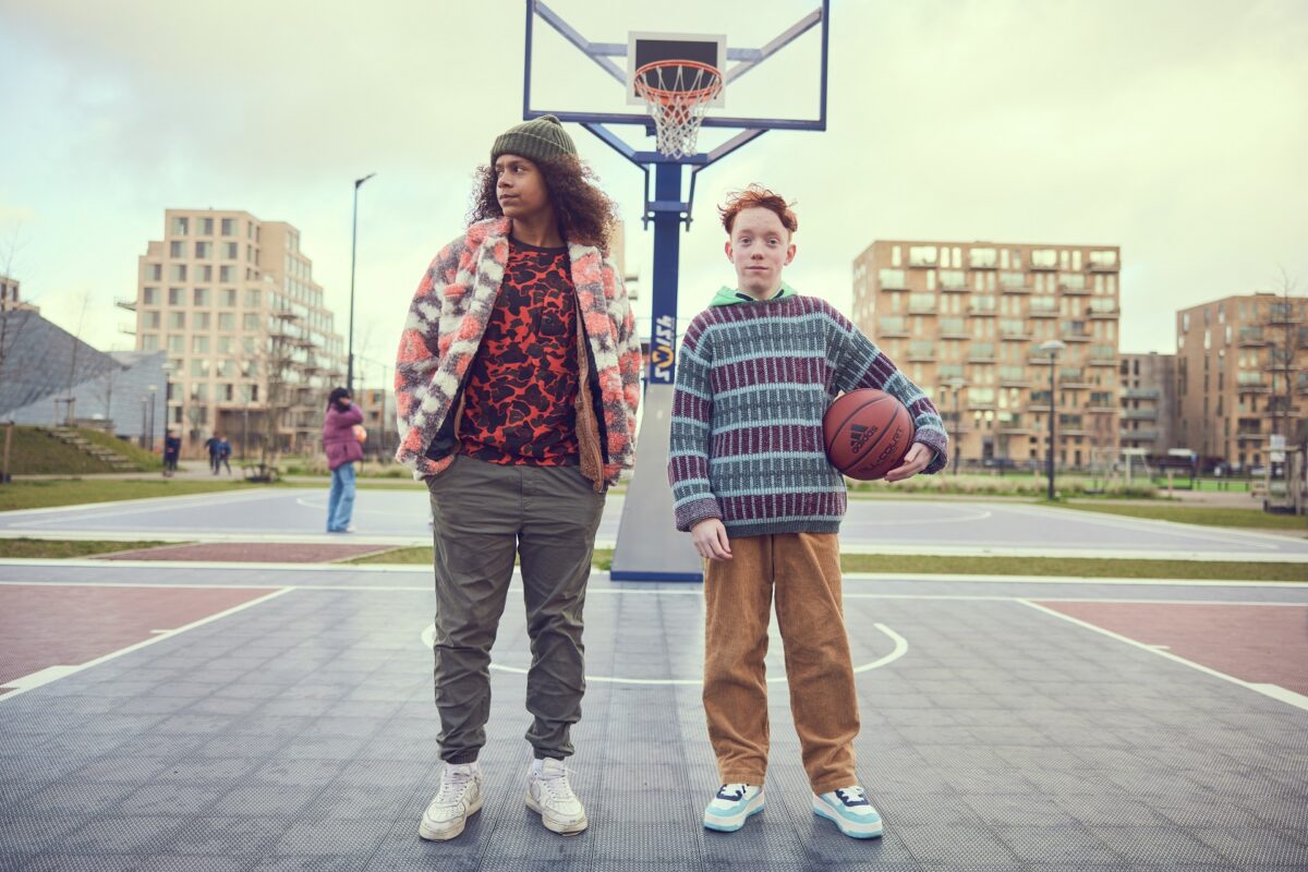 Twee jongens staan op een rookvrij schoolplein naast elkaar. De jongen links kijkt naar de zijkant en de jongen links kijk recht in het beeld en heeft een basketbal in zijn handen.