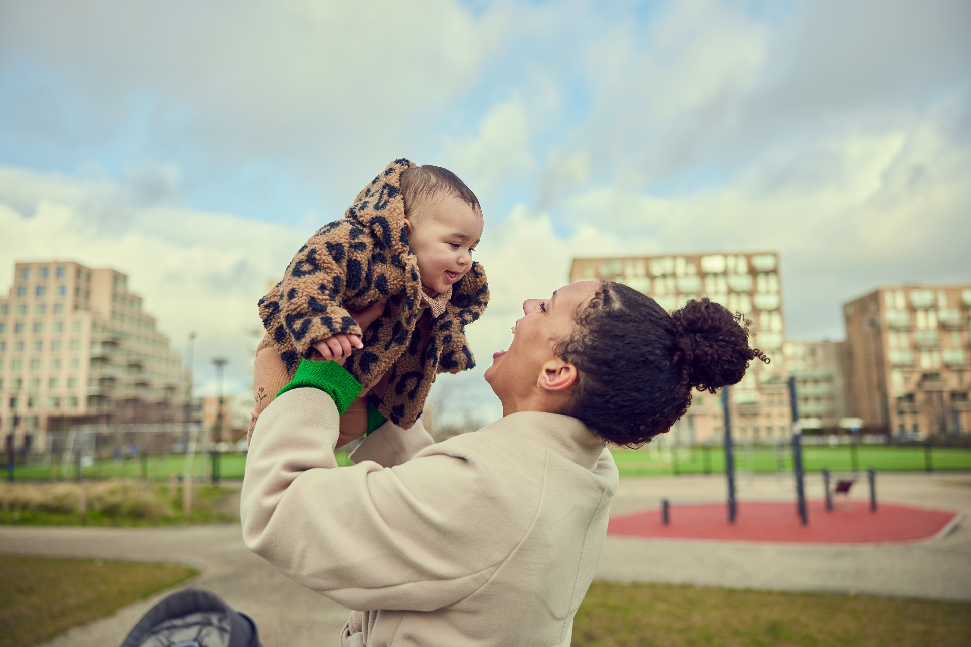 Baby wordt vastgehouden door moeder. Ze zijn op een rookvrij schoolplein en kijken elkaar vrolijk aan.